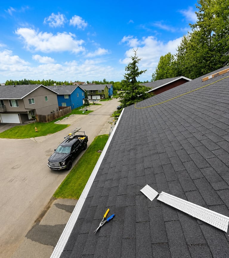 Aerial view of a residential street with asphalt roofing in foreground, houses, parked car, and green trees under blue sky