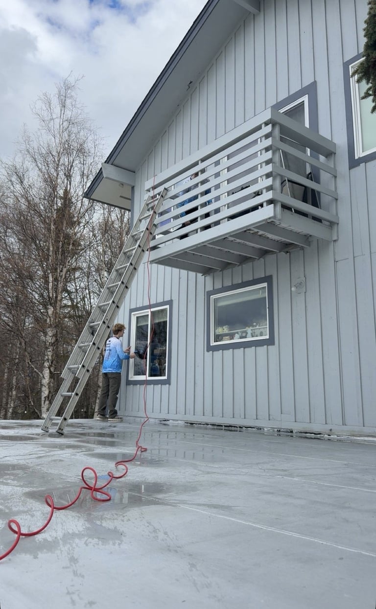 Person on ladder cleaning metal sided house exterior with power washer and red hose on wet concrete patio
