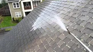 A residential roof being pressure washed, showing water spray cleaning the shingle surface